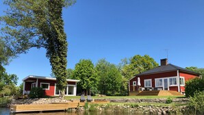 Una cabaña roja con un muelle que se adentra en un lago tranquilo, rodeada de exuberante vegetación y árboles.