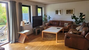 Olohuoneessa on ruskea nahkasohva, litteänäyttöinen televisio, sohvapöytä ja pieni sivupöytä. Huoneessa on kaksi ikkunaa, joissa on verhot, sekä ruukussa oleva kasvi. Ulkona näkyy myös polkupyörä.