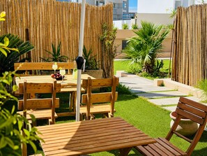 Een houten eethoek buiten, met een tafel en stoelen, een bamboebeschoeiing en een kleine tuin met palmstruiken.