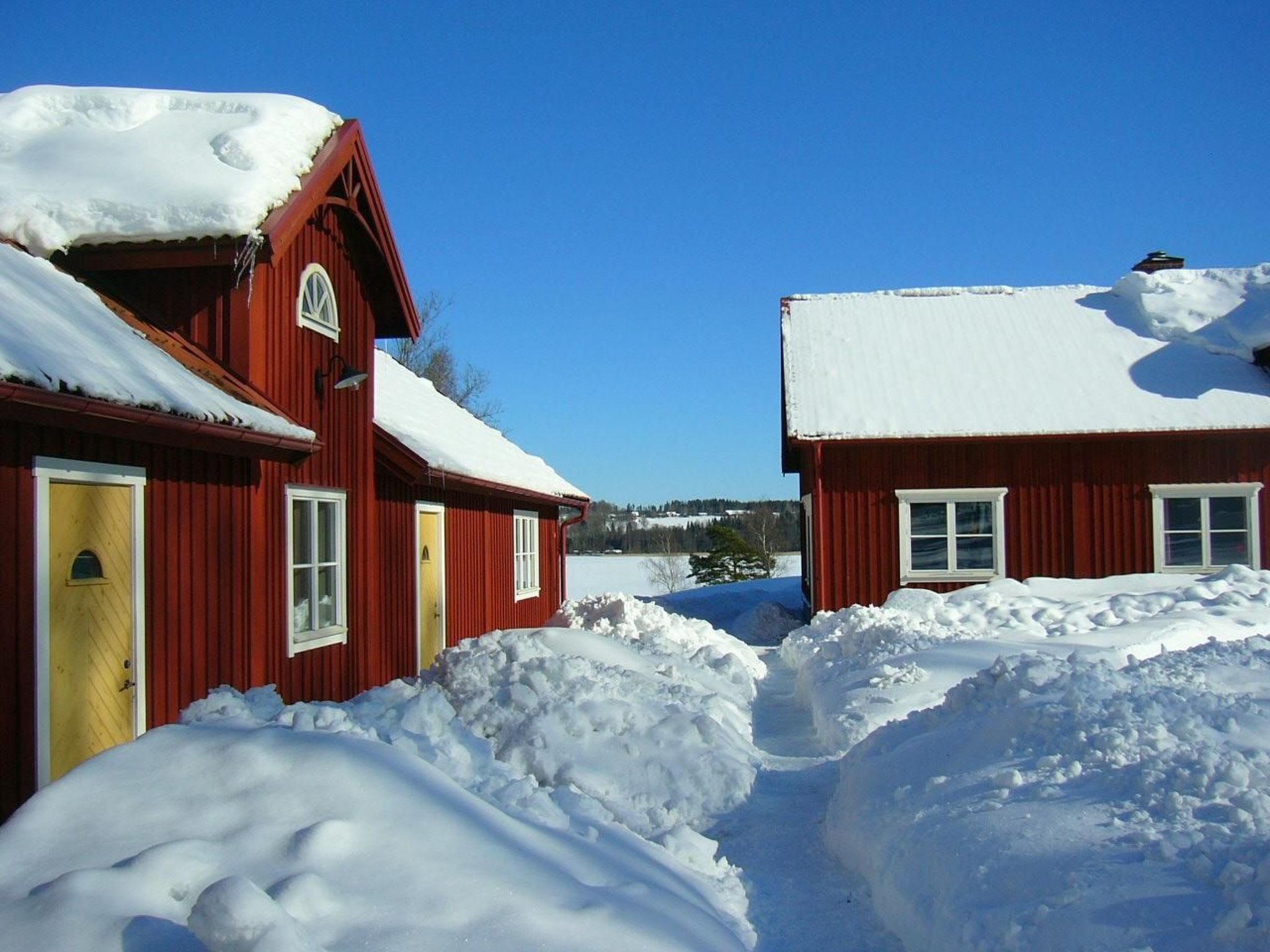 Deux maisons en bois rouges, avec des toits couverts de neige, sous un ciel bleu clair.