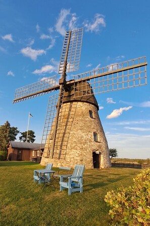 Een stenen windmolen met een grote houten constructie, twee blauwe stoelen en een grasveld.