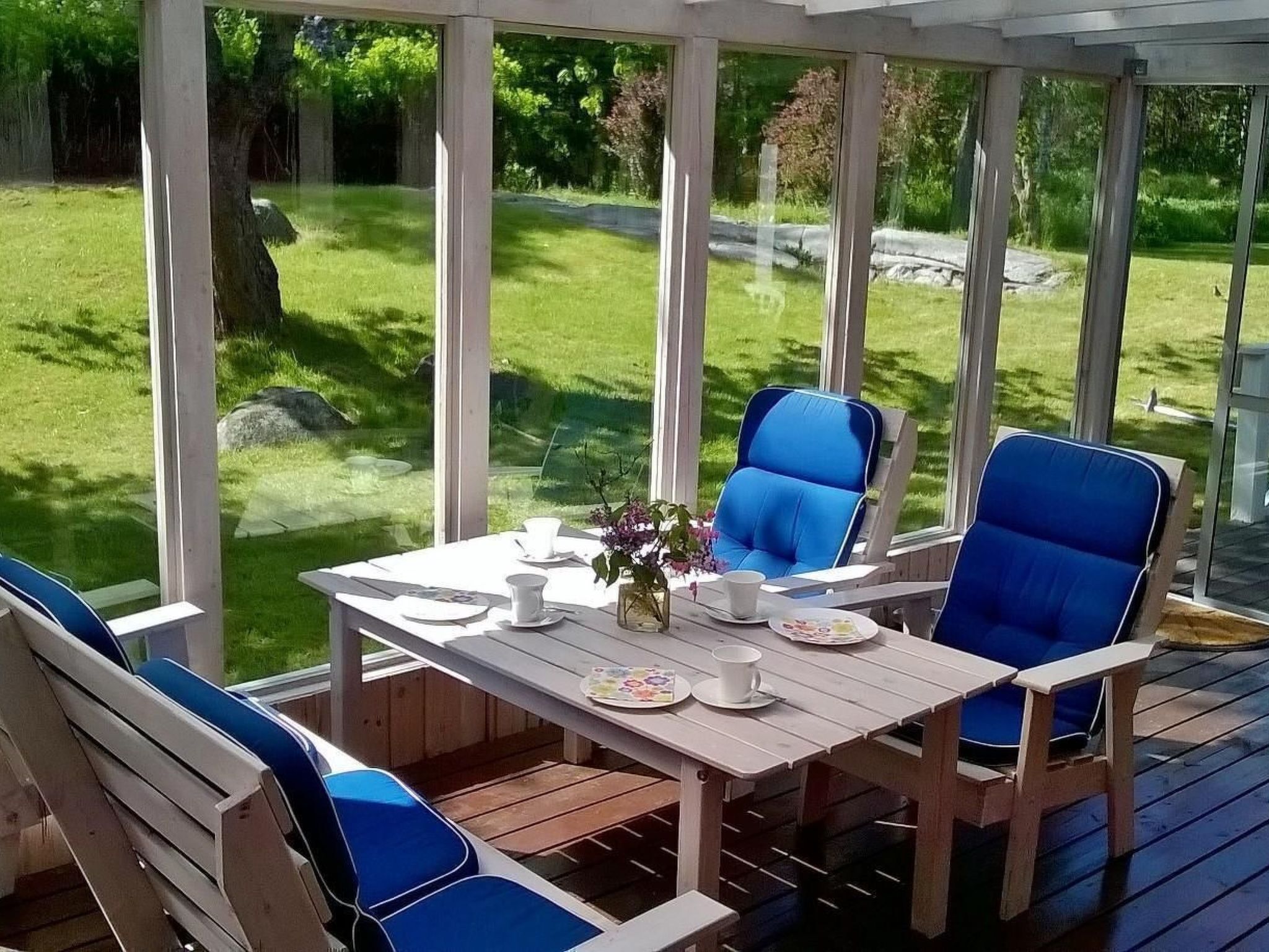 Eine überdachte Terrasse mit einem Holztisch und blauen Stühlen, mit Blick auf eine grüne Fläche mit Bäumen.