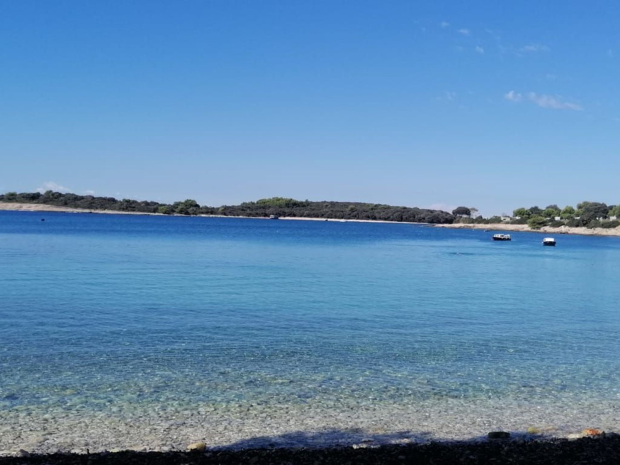 A clear view of a calm sea with boats, a sandy beach, and a distant shoreline with vegetation.