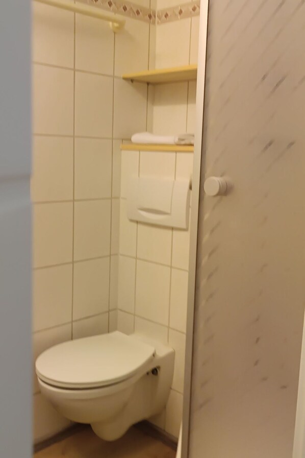 A small, basic bathroom with a toilet, a shelf above the toilet, and a towel hanging on the wall.