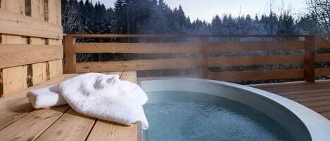 A wooden deck with a hot tub, surrounded by a wooden fence and snow-covered trees.