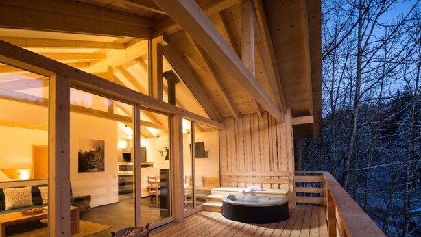 En træterrasse med jacuzzi og siddeplads, store glasdøre der fører til et indendørs rum, samt udsigt over et skovområde.