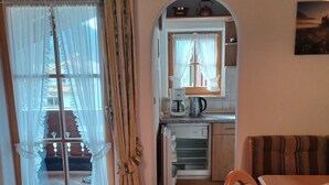 Un salon chaleureux avec un plafond en bois, une table à manger et un canapé. On aperçoit une cuisine à travers une arche surmontée d’une fenêtre, et un escalier mène à l’étage supérieur.