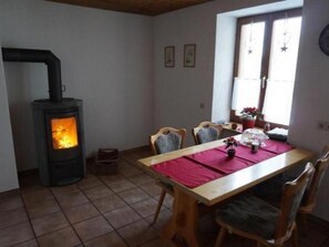 Ein Essbereich mit Tisch für vier Personen, einem Kaminofen und einem Fenster mit Vorhängen.