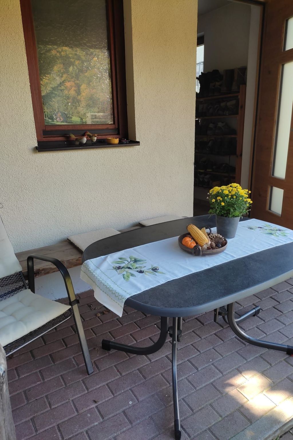 An outdoor patio with a table set for two, featuring a tablecloth, a potted plant, and a bowl of corn and other vegetables.