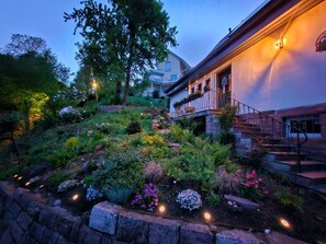 Um jardim com diversas plantas e flores, um muro de pedra e um edifício com escadaria.