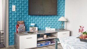 Un salon moderne avec un mur bleu à texture, un téléviseur à écran plat fixé au-dessus d’un meuble de rangement en bois, et une pancarte indiquant « Bienvenue ».