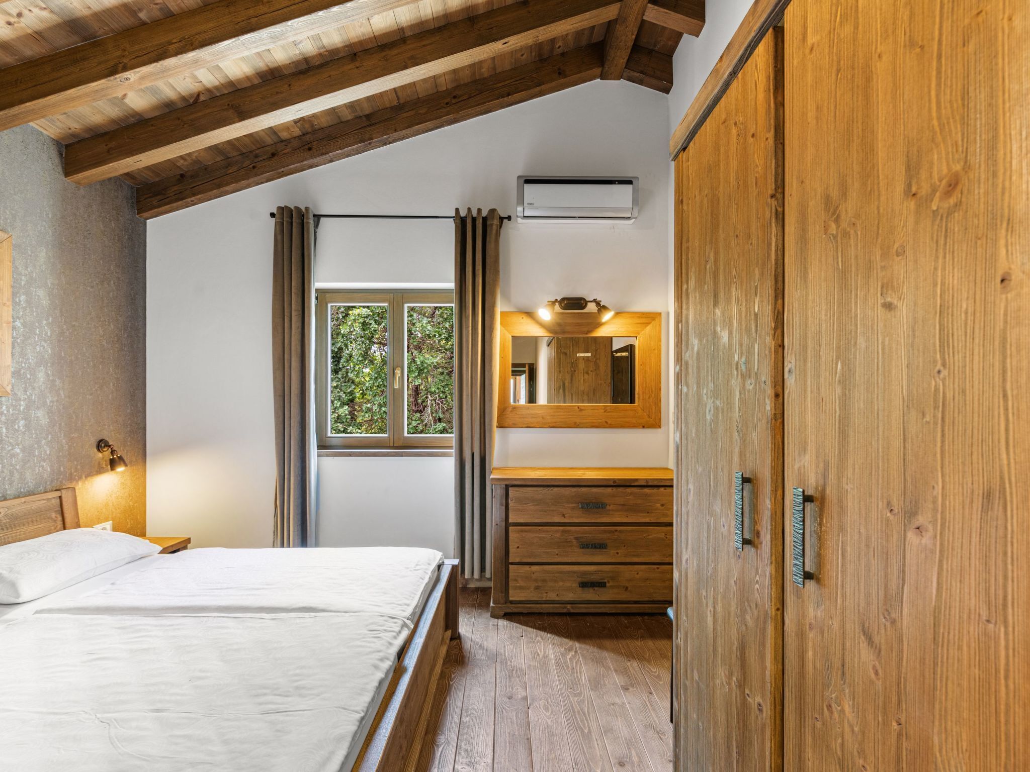 A bedroom with a wooden ceiling, a bed, a nightstand, a dresser, a mirror, and an air conditioning unit.