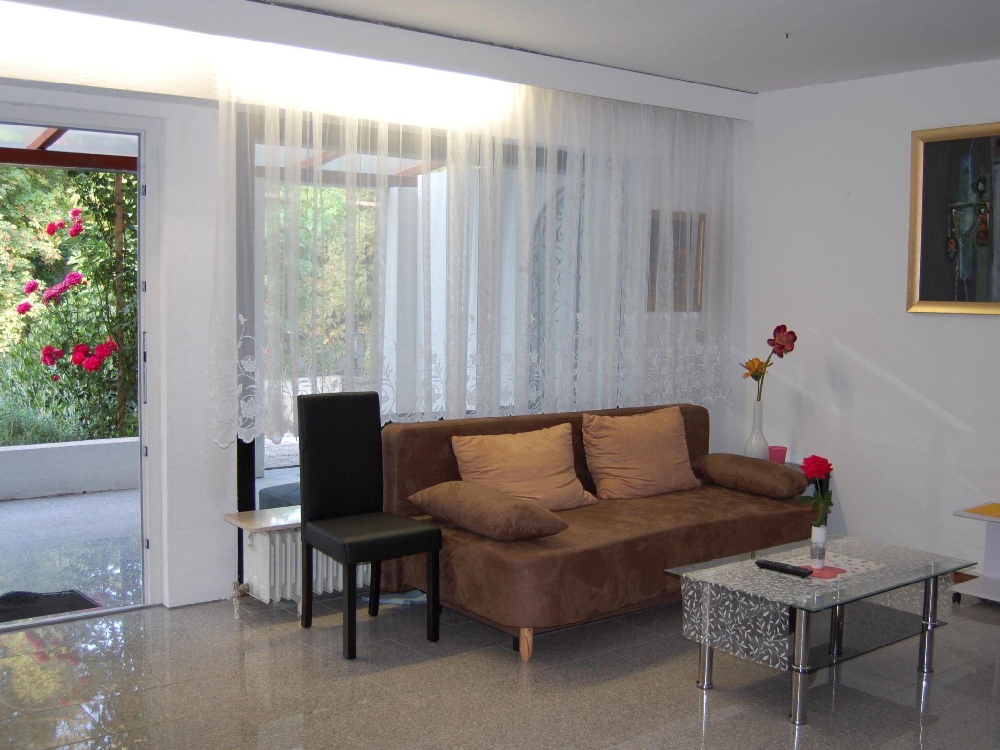 Een moderne woonkamer met een bruine bank, een glazen salontafel en uitzicht op groen door dunne gordijnen.