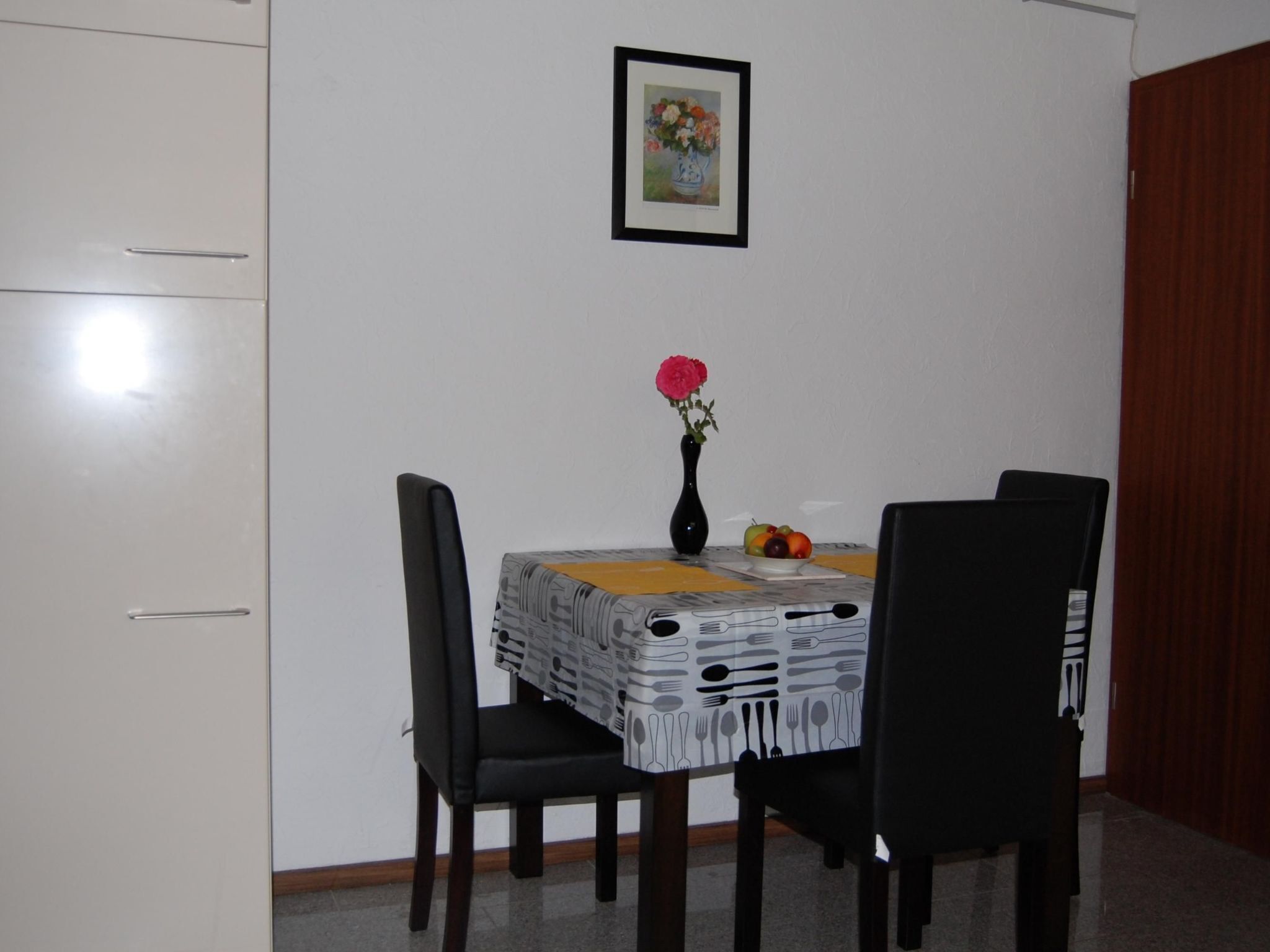 A dining area with a table set for two, a vase with flowers, and a framed picture on the wall.