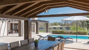Um pátio externo com mesa e cadeiras de madeira, uma piscina e um pérgola.