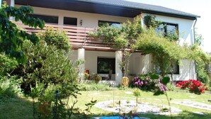 Et moderne hus i to etager med en overdækket terrasse, omgivet af frodig vegetation og en lille fiskedam.