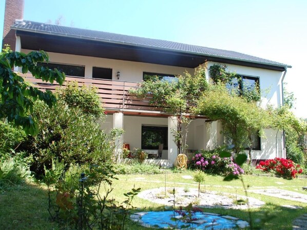 Een modern, twee verdiepingen tellend huis met een overdekte patio, omgeven door weelderige begroeiing en een kleine vijver.