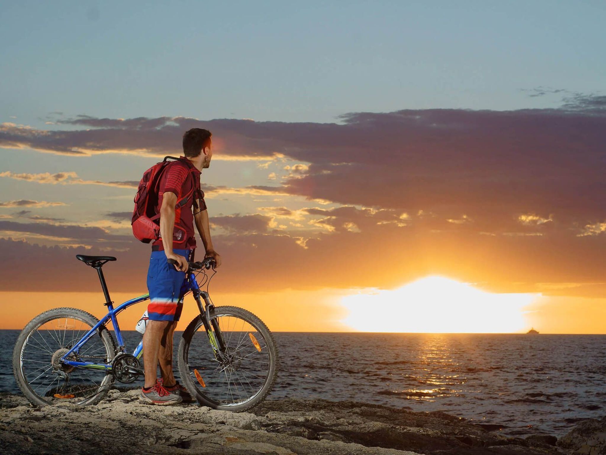 Eine Person steht neben einem Fahrrad an einem felsigen Ufer, im Hintergrund ein Sonnenuntergang.