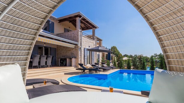 Ein Poolbereich mit Liegestühlen und Sonnenschirmen, ein Gebäude mit Steinbau und einem Balkon sowie ein klarer, blauer Himmel.