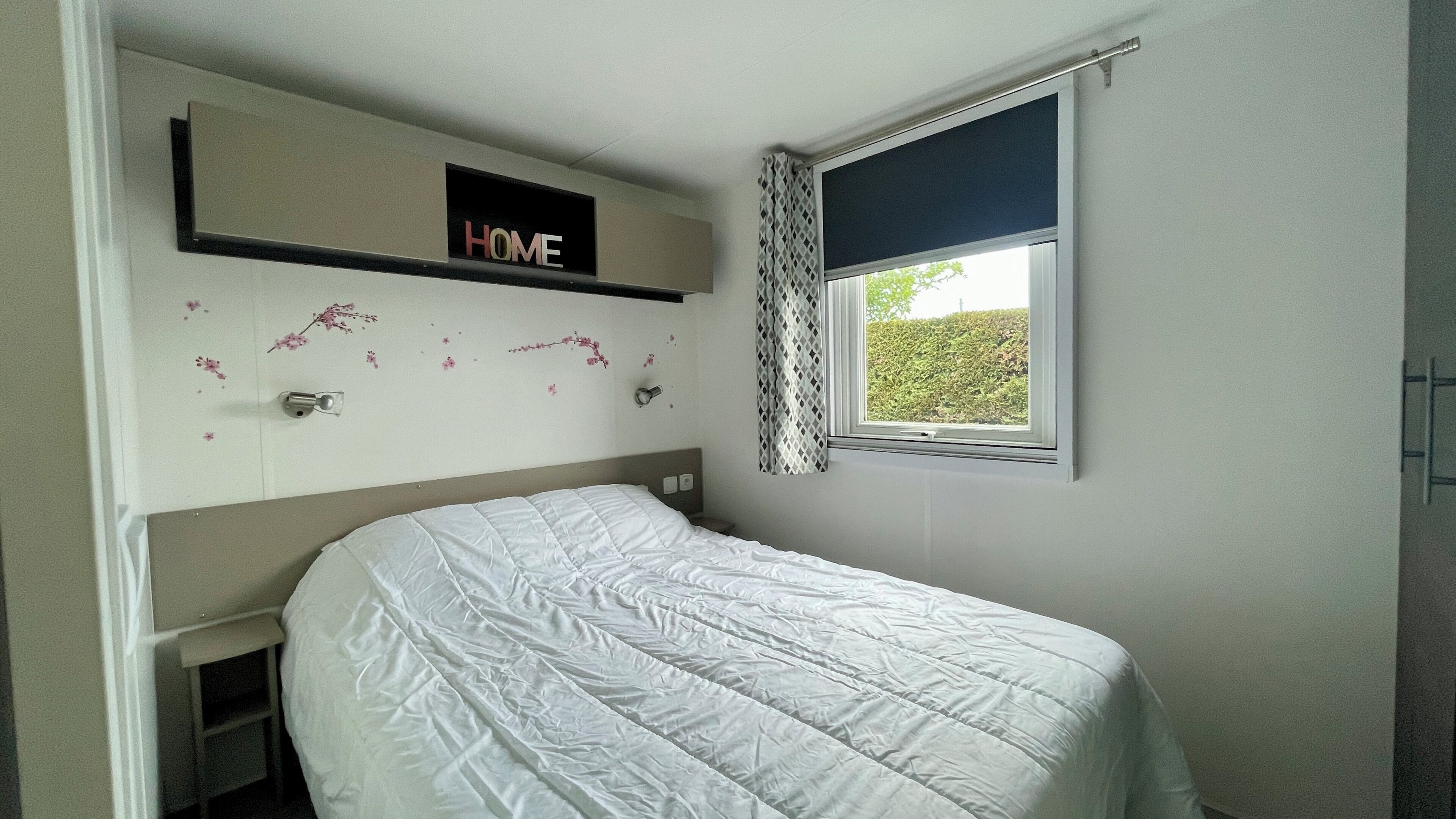 A bedroom with a bed, a 'HOME' sign, a window with a view, and a wall-mounted shelf.