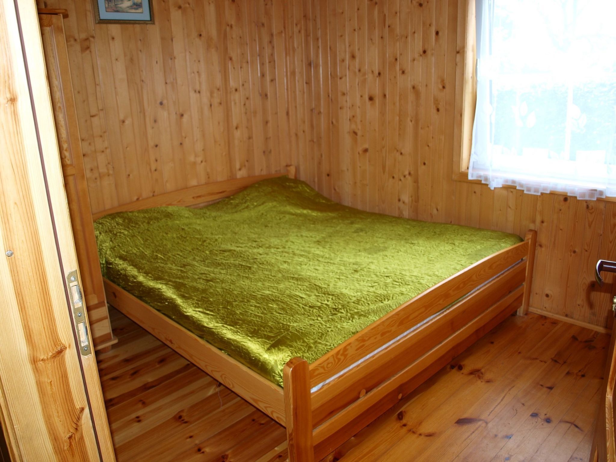 Ein Zimmer mit Holzboden, einem Einzelbett, einem Fenster mit weißem Vorhang und einer Holztür.