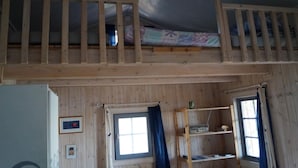 Un dormitorio tipo loft con una cama nido de madera, dos ventanas con cortinas, un estante con objetos y un cuadro en la pared.