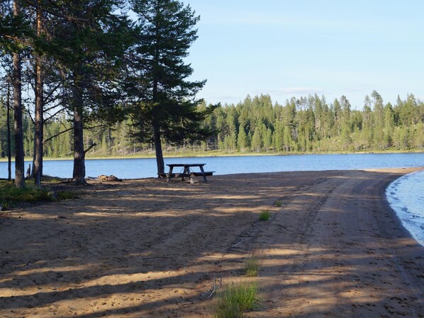 Une plage de sable avec une table de pique-nique et un banc, entourée d’arbres et bordée par un lac.