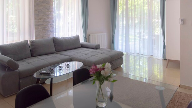 Ein modernes Wohnzimmer mit einem grauen Ecksofa, einem Glastisch und einer Vase mit Blumen.