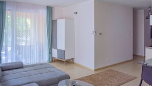 Ein modernes Wohnzimmer mit grauem Sofa, einem Glastisch und einem beigen Teppich. Es gibt einen grauen Kleiderschrank, einen kleinen Tisch mit Lampe sowie ein Fenster mit leichtem Vorhang.