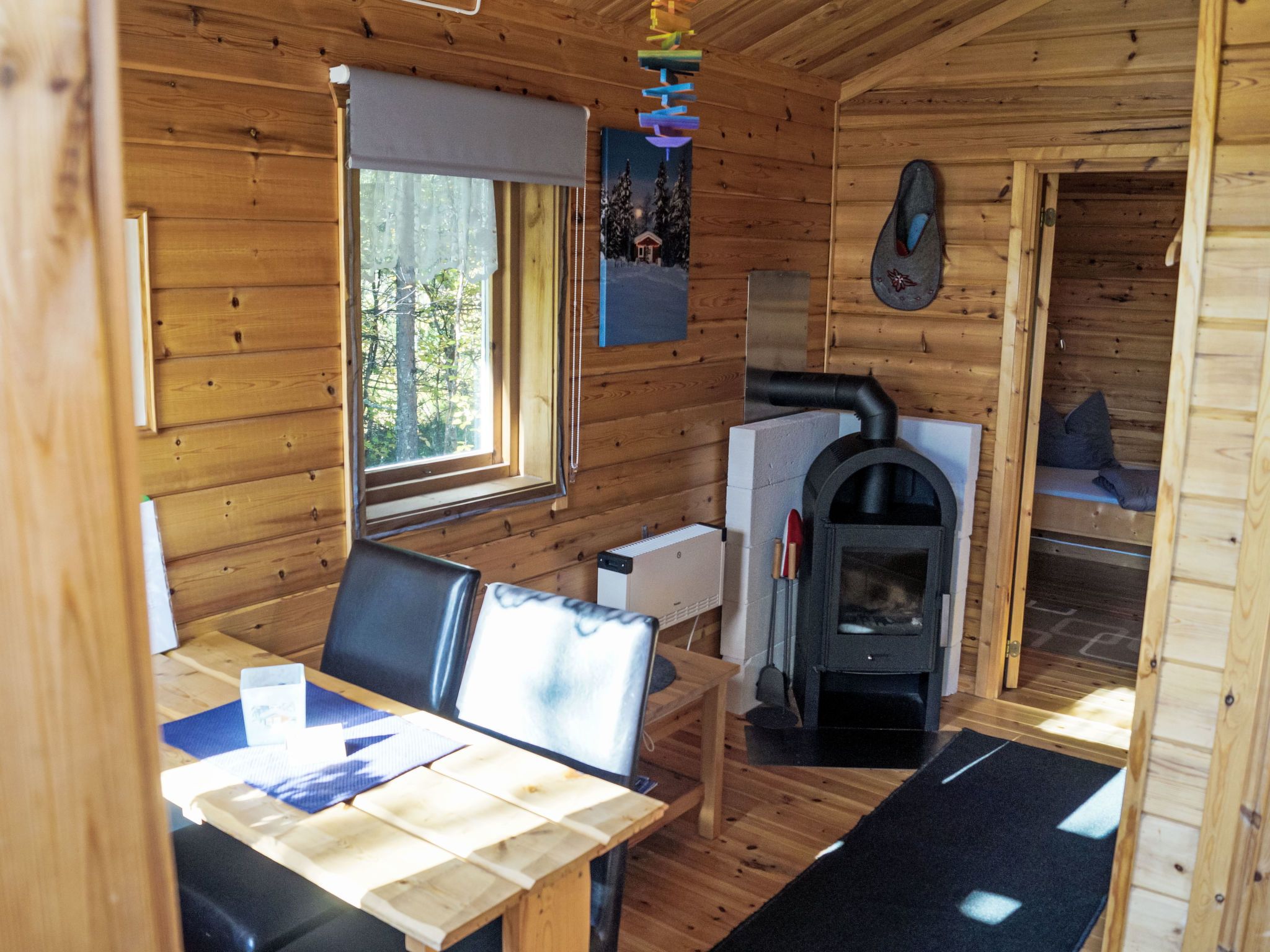 A wooden cabin interior with a dining table, chairs, a fireplace, and a window with curtains.