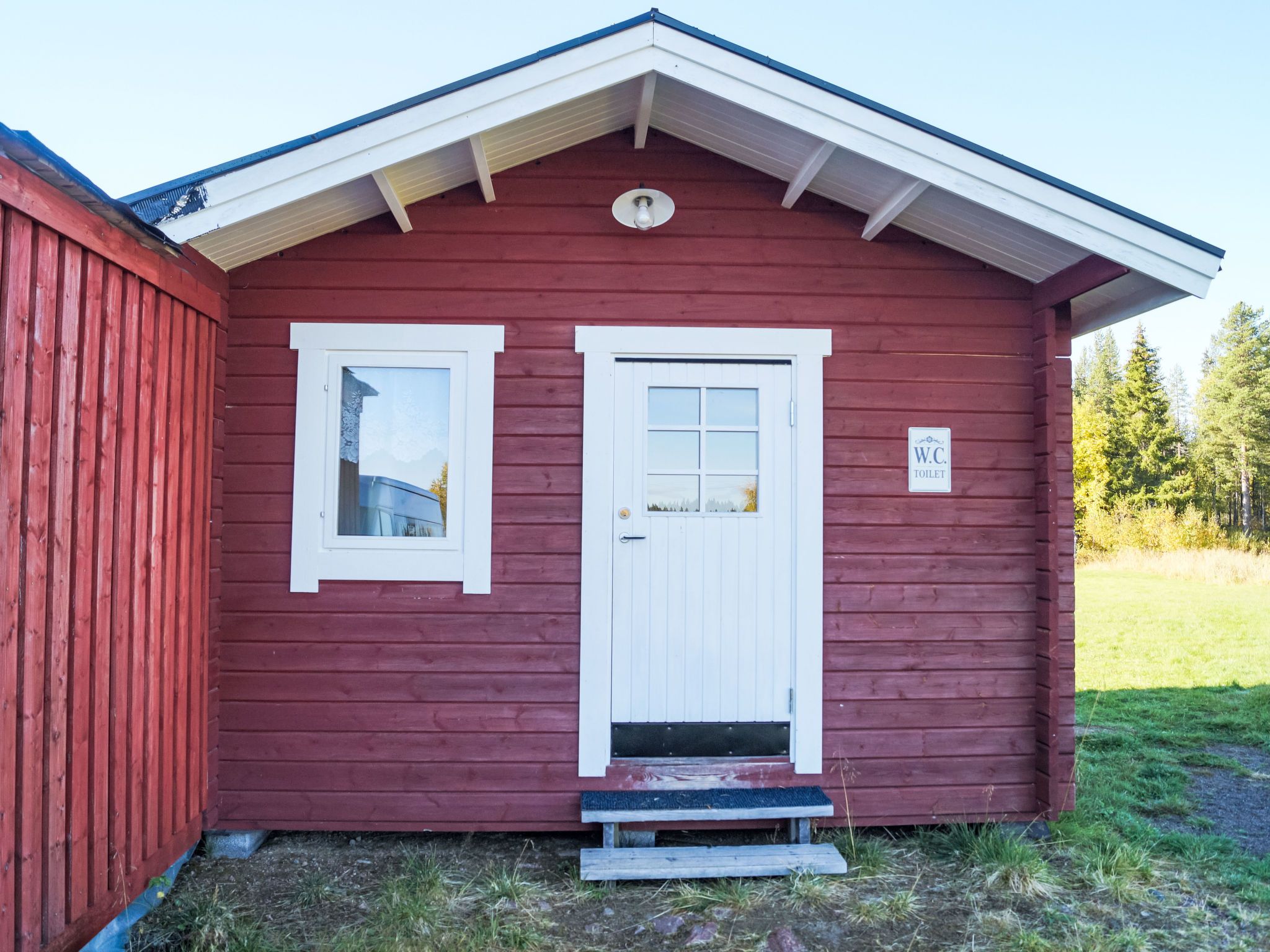 A red wooden building with a white door and window, a small porch, and a sign indicating a toilet.