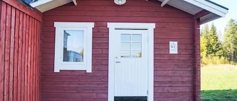 A red wooden building with a white door and window, a small porch, and a sign indicating a toilet.