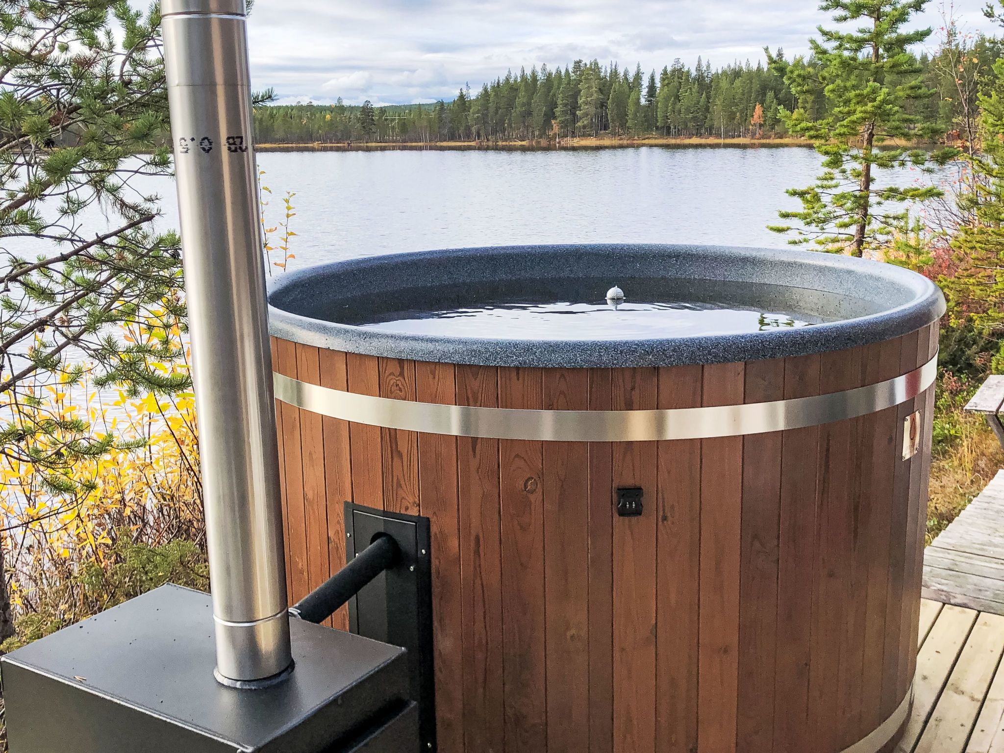 En bubbelpool med träpanel på utsidan och en metallram, placerad på ett trädäck med utsikt över en sjö och träd i bakgrunden.