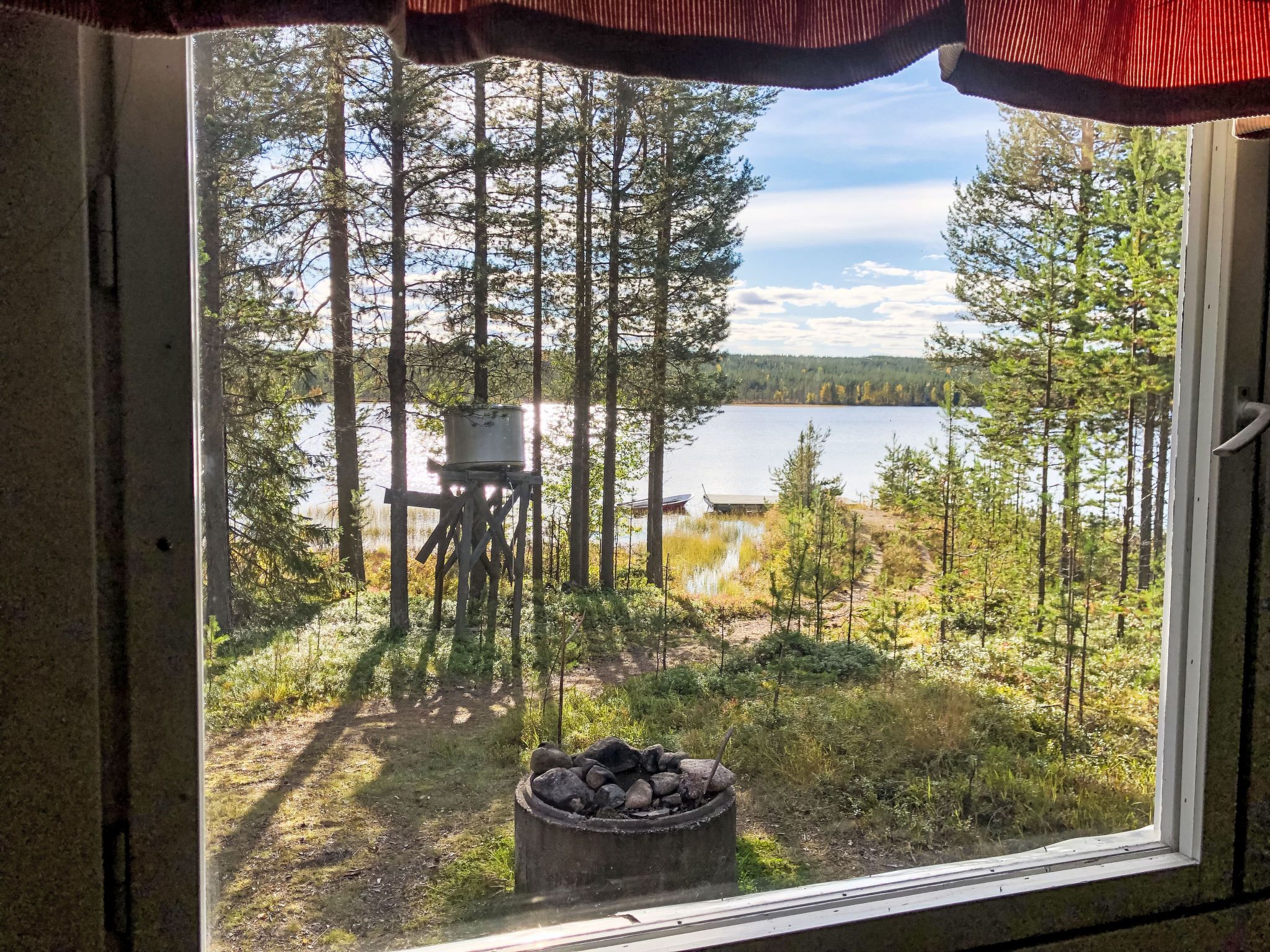 A window view of a forested area with a lake, a fire pit, and a deck chair.