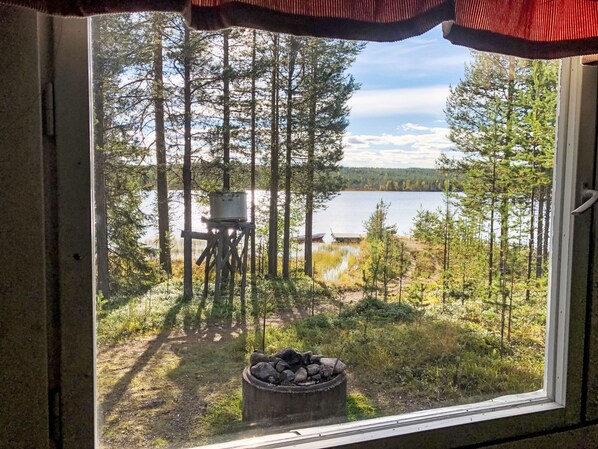 A window view of a forested area with a lake, a fire pit, and a deck chair.