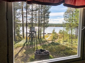 A window view of a forested area with a lake, a fire pit, and a deck chair. - in Arvidsjaur mit eigener Sauna by Interhome (Arvidsjaur)