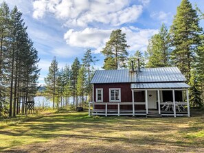 Uma cabana com telhado de metal, varanda branca e vista para um lago rodeado de árvores.
