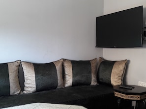 A black sofa with beige and dark cushions, a small side table with a remote control, and a mounted TV on the wall. - Résidence Mogador 1 by Interhome (Agadir)
