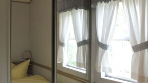 Una camera da letto con un ampio specchio, due finestre con tende, un letto e un comodino con una lampada.