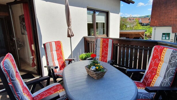 An outdoor patio with a round table, four chairs with patterned cushions, a wicker basket with flowers, and a view of a building with a sloped roof. - Casa Maria in Ehrenberg an der Rhön by Interhome (Ehrenberg)