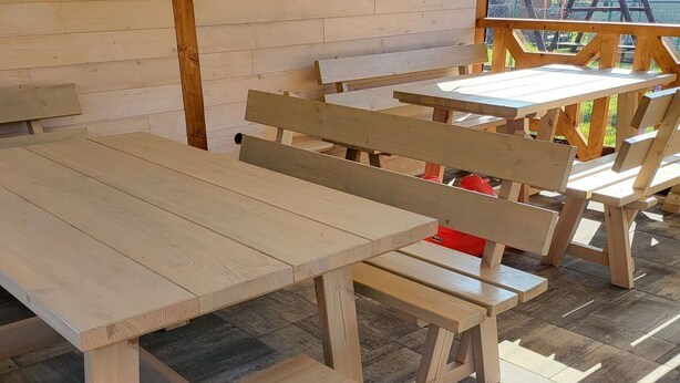 Un espace extérieur abrité, doté de bancs et de tables en bois, d’un sol carrelé et offrant une vue sur un bâtiment à travers une fenêtre.