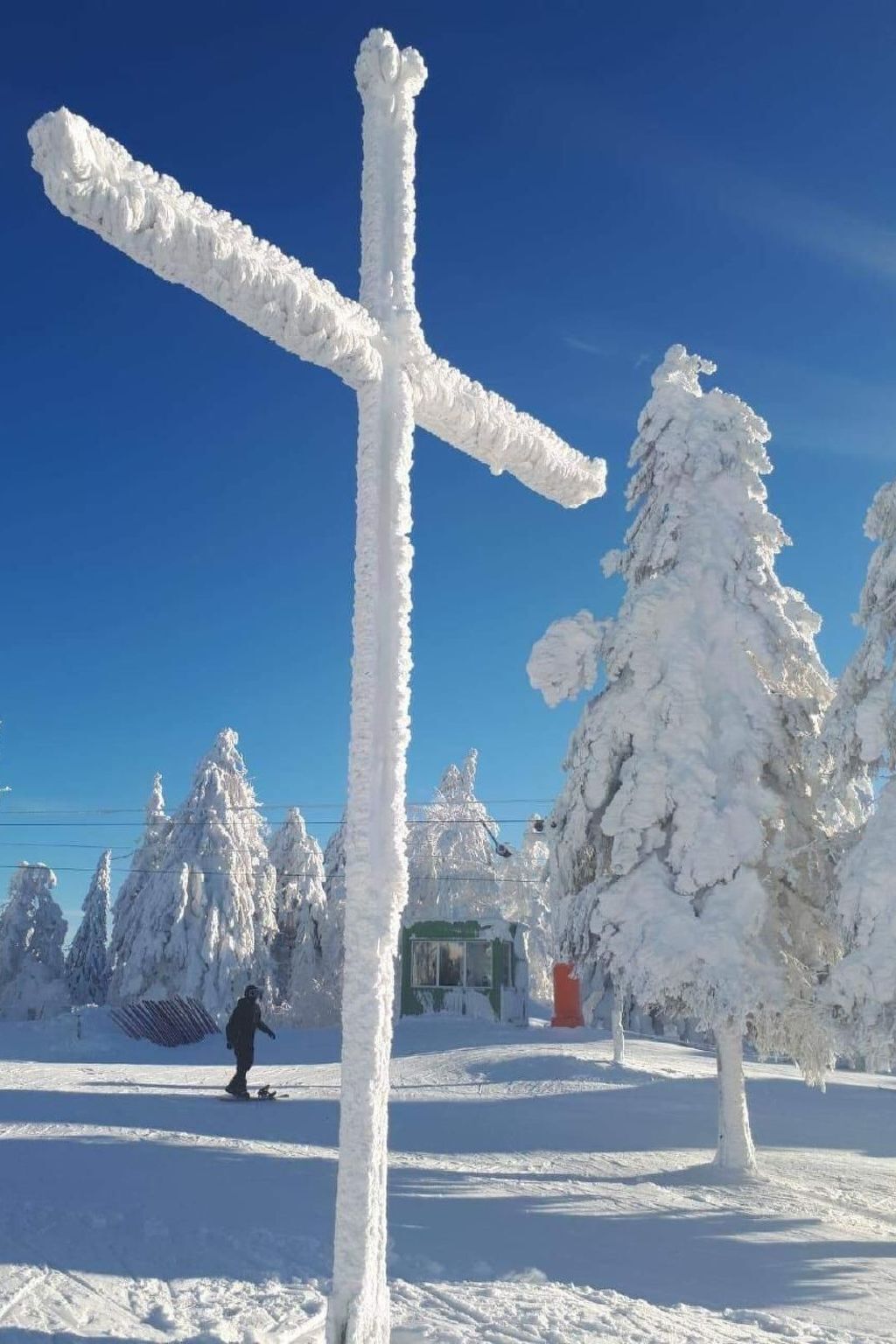 A snow-covered cross in a snowy landscape with trees and a clear blue sky.