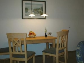 A dining area with a wooden table, two chairs, a basket of fruit, a bottle of wine, and a framed picture on the wall. - Am Buchenhain in Koserow by Interhome (Koserow)