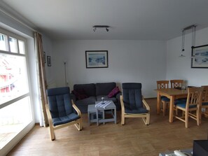 A living room with a sofa, two armchairs, a coffee table, and a dining table with chairs. There is a window with curtains, a framed picture on the wall, and a ceiling light fixture. - Am Buchenhain in Koserow by Interhome (Koserow)
