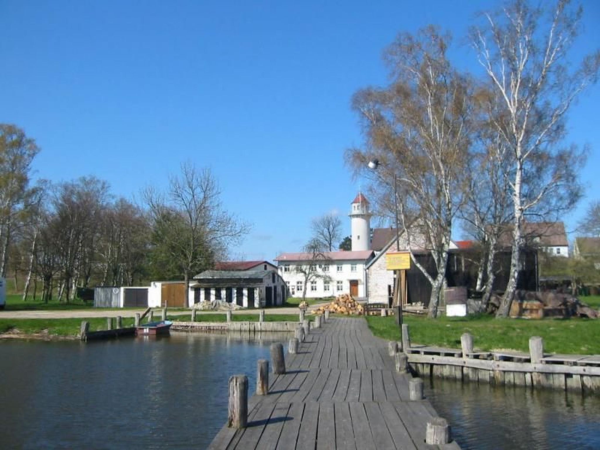 Un muelle de madera que conduce a un edificio blanco con techo rojo, un faro al fondo y un río tranquilo.