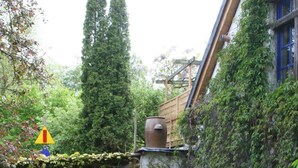 Uma trilha no jardim conduz a um edifício rústico com uma parede de pedra, um grande vaso de barro e uma estrutura de madeira no telhado.