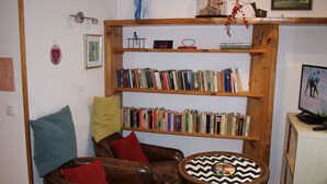 Uma sala de estar aconchegante com uma estante de madeira repleta de livros, uma poltrona de couro confortável com almofadas vermelhas, uma pequena mesa de madeira com tampo estampado e uma televisão sobre um suporte.