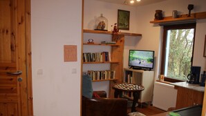 Uma sala de estar acolhedora com móveis de madeira, uma estante de livros, uma televisão e uma janela com vista para árvores.
