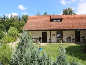 Een gebouw met twee verdiepingen, voorzien van een dak met rode tegels, een overdekte veranda met buitenmeubilair en een verzorgde tuin met diverse planten.