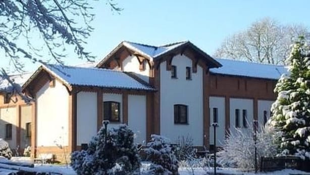 Ein mit Schnee bedecktes Gebäude mit mehreren Fenstern und einem geneigten Dach, umgeben von schneebedeckten Bäumen und Sträuchern.